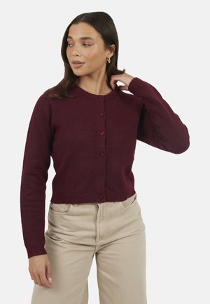 Kobieta w bordowym swetrze na guziki i beżowych spodniach, poprawiająca włosy jedną ręką, spoglądająca w bok na białym tle.