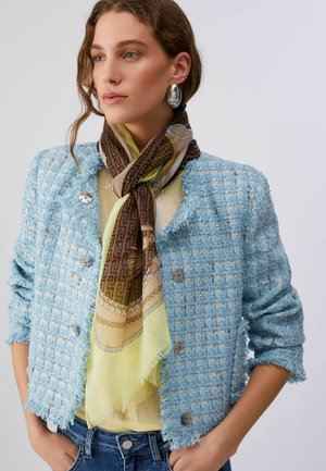 Donna con giacca azzurra texturizzata, sciarpa marrone e gialla a motivi, orecchino argentato di grande impatto, con le mani nelle tasche dei jeans.