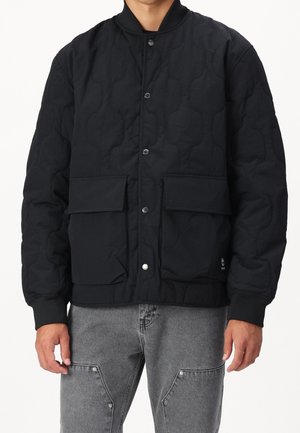 Hombre con chaqueta negra acolchada con botones a presión delanteros y dos grandes bolsillos con solapa, combinada con vaqueros grises con remaches en los bolsillos.