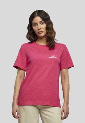 Femme aux cheveux ondulés foncés portant un t-shirt rose vif avec un texte blanc "THINK DIFFERENT." et un pantalon beige, debout devant un fond gris uni.