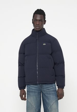 Marineblå pufferjakke med høj krave, lynlåslukning og Lacoste-logo. Har kviltede paneler og elastiske manchetter. Bæres med lyse denimjeans.