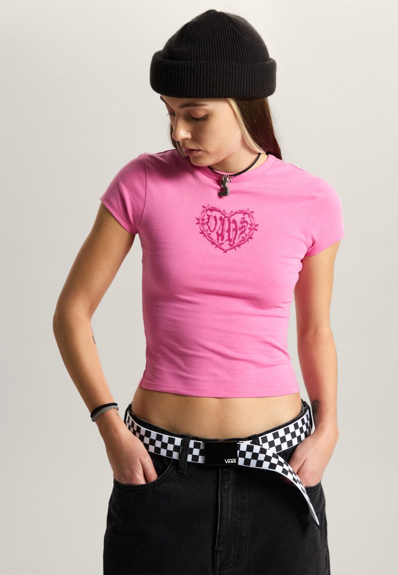 Joven con gorro negro, camiseta ajustada rosa con gráfico de corazón, cinturón de cuadros de la marca Vans y pantalones negros, con las manos en los bolsillos, mirando hacia abajo.