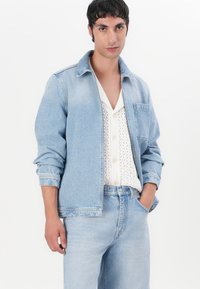 Lys blå denimjakke med lange ermer, frontlomme og en avslappet passform. Båret over en hvit genser med strukturert strikk og knapper.