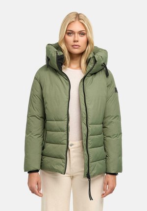 Femme blonde portant une veste matelassée verte avec capuche, un pantalon beige et un haut beige clair, debout devant un fond uni.