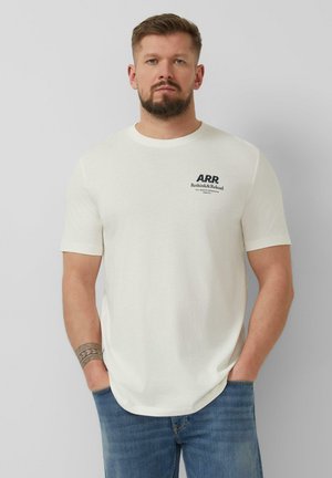 Mann mit Bart, der ein weißes A.R.R. Rethink & Reload T-Shirt und blaue Jeans trägt, mit den Händen in den Taschen stehend vor einem schlichten Hintergrund.