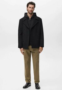 Cappotto peacoat nero con doppiopetto e grandi revers, indossato sopra un hoodie nero con zip, abbinato a pantaloni beige e stivali neri.
