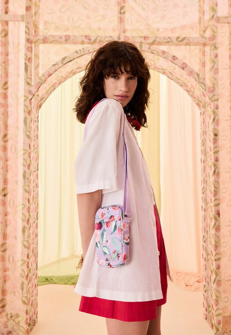 Femme aux cheveux bouclés et foncés portant une chemise blanche ample et une jupe rouge, portant un petit sac bandoulière violet à motif floral.