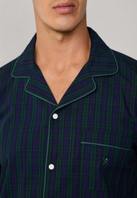 Camisa de pijama de manga corta a cuadros verdes y marinos con botones blancos, bordes ribeteados y un bolsillo en el pecho que presenta un logo sutil.