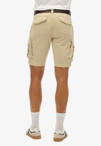 Pantalones cortos de cargo beige con bolsillos laterales y solapas traseras, hechos de una tela ligera. Llevados con calcetines blancos y zapatillas casuales.