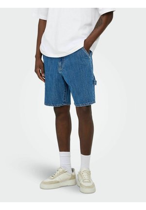 Osoba nosząca niebieskie jeansowe szorty do połowy uda, białe skarpetki typu crew, kremowe trampki i białą koszulę z krótkim rękawem, stojąca z jedną ręką w kieszeni.