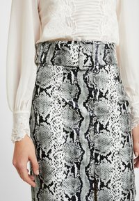 Närbild av händer som bär en vit genomskinlig blus med blommig broderi och en högmidjad kjol i grått och svart ormskinnsmönster med ett bälte.