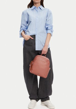 Mujer con una camisa azul claro abotonada y jeans negros holgados, sosteniendo una pequeña mochila rosa con zapatillas blancas sobre un fondo blanco.