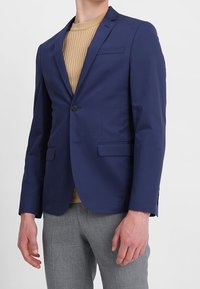 Man som bär en marinblå kavaj över en beige ribbad tröja och grå strukturerade byxor, står mot en enkel ljus bakgrund.