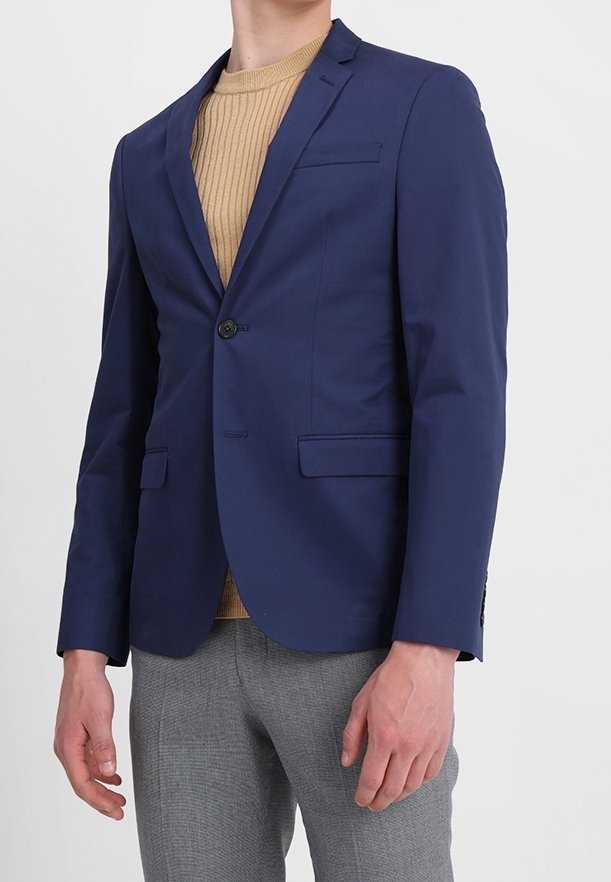 Man som bär en marinblå kavaj över en beige ribbad tröja och grå strukturerade byxor, står mot en enkel ljus bakgrund.