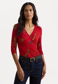 Rød innpakningsbluse med trekvart ermer, prydet med gullkjede og solmønstre. Kombinert med mørke jeans og et svart belte med gullspenne.