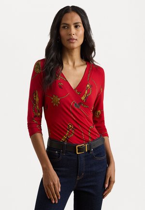Red wrap top with three-quarter sleeves, featuring gold chain and sun patterns. Paired with dark jeans and a black belt with a gold buckle.