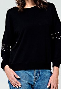 Felpa nera con dettagli in pizzo e perle sulle maniche, caratterizzata da una vestibilità ampia e orlo a coste, abbinata a jeans in denim blu chiaro.