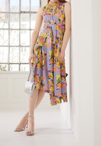 Robe à motif floral aux nuances de violet, jaune et vert, dotée d'une taille à nouer et d'un ourlet asymétrique. Sac à main argenté et talons à lanières.