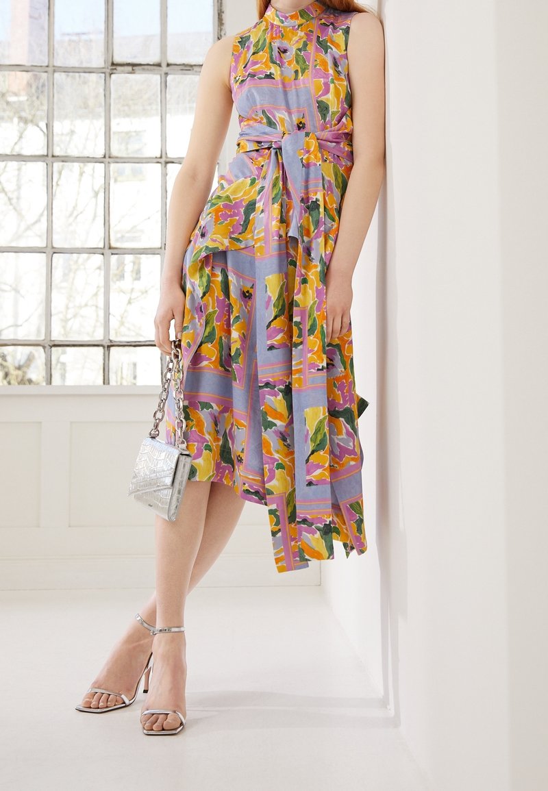 Robe à motif floral aux nuances de violet, jaune et vert, dotée d'une taille à nouer et d'un ourlet asymétrique. Sac à main argenté et talons à lanières.