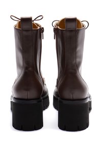 Ein Paar braune Leder-Stiefeletten mit derben schwarzen Absätzen und seitlichen Reißverschlüssen, von hinten auf einem weißen Hintergrund gezeigt.