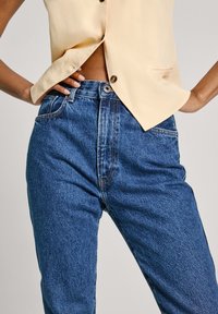 Ψηλοκάβαλο μπλε τζιν παντελόνι με κλασική εφαρμογή, που διαθέτει απαλή φθορά, ορατές ραφές και κλείσιμο με έναν μόνο κουμπί.