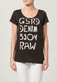 Woman wearing a black short-sleeve shirt with white text "GSRD DENIM 5204 RAW" and white pants, standing against a light background.