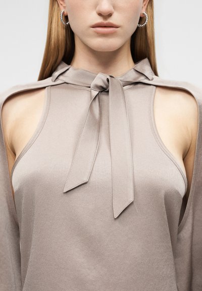 Femme portant un haut sans manches taupe avec un col noué en forme de nœud et des épaules découpées, assorti à des boucles d'oreilles anneaux argentées, sur un fond uni.