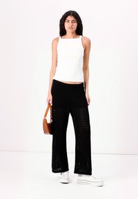 Top sin mangas blanco con relieve, pantalones de pierna ancha negros de punto y zapatillas blancas. Un bolso marrón cuelga del hombro.