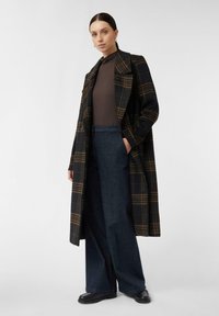Doppelreihiger schwarzer und brauner karierter Übermantel, weite blaue Denim-Hosen, dazu ein hochgeschlossener brauner Oberteil und schwarze Schuhe.