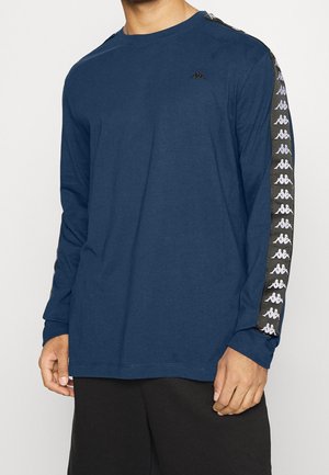 Mann trägt ein dunkelblaues Langarmshirt mit schwarz-weißem Logoband entlang der Ärmel und schwarze Hose.