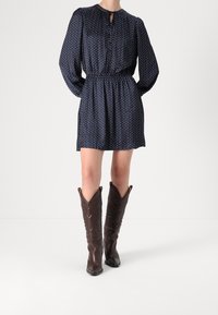 Nainen, jolla on yllään tummansininen pilkullinen pitkähihainen mekko, jonka vyötärö on kerätty, sekä polven korkeiset tummanruskeat cowboy-kengät, seisoo valkoisella lattialla.