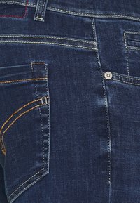 Jeans de mezclilla azul oscuro con múltiples bolsillos, costuras contrastantes en amarillo y rojo, y detalles en metal plateado. Textura suave.