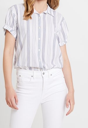 Chemise à manches courtes, boutonnée, de couleur blanche avec de fines rayures verticales bleues et grises. Portée avec un jean blanc, affichant une coupe décontractée.