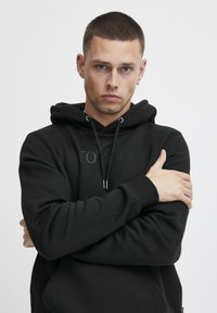 Black hoodie with drawstring, kangaroo pocket, and embossed "TOKYO" text. Model stands with arms crossed against a white background.