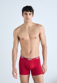 Rote Boxerbriefs mit einem kontrastierenden schwarzen Bund, auf dem der Markenname "BOSS" steht. Der Stoff wirkt glatt und eng anliegend.