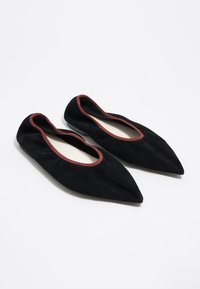 Zapatos planos de ante negro con punta afilada y ribete rojo a lo largo de la abertura, presentando una superficie suave y texturizada y un diseño minimalista.