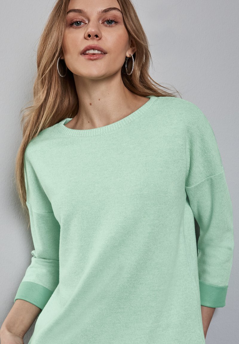 Ljus mintgrön tröja gjord av mjukt stickat material. Har en rundad halslinning och 3/4 ärmar med matchande manchetter. Slät yta och avslappnad passform.