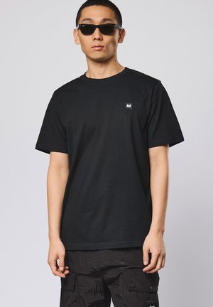 Ung mann med svarte solbriller, en ensfarget svart t-skjorte med en liten logo, og svarte cargobukser foran en grå bakgrunn.