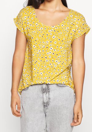 Kvinde iført gul kortærmet bluse med hvidt blomstertryk og lysegrå højtaljede jeans, står med afslappede arme.