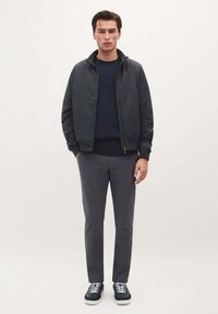 Mørk bomberjakke over en marineblå sweatshirt, parret med grå bukser og mørke sneakers. Glat stof og minimalistisk design hele vejen igennem.