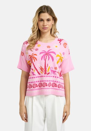 Woman wearing a pink short-sleeve sweater with colorful palm tree, flower, and animal prints paired with white trousers.
