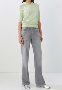 Haut tricot vert clair à manches courtes, associé à un jean gris évasé. Des chaussures plates argentées complètent la tenue. Texture lisse, design simple.