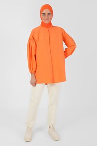 Blusa arancione brillante a maniche lunghe con polsini arricciati, chiusura con bottoni e colletto alto, abbinata a pantaloni bianchi a gamba dritta e sneakers beige.