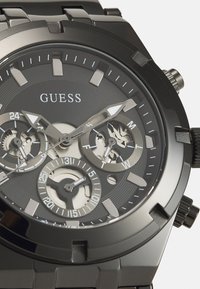 Reloj cronógrafo de acero negro con una esfera texturizada negra, acentos plateados y múltiples subesferas que muestran el día y la hora en formato de 24 horas.