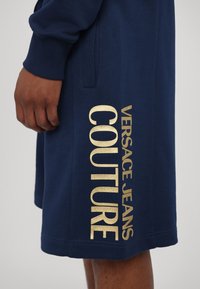 Temno modra kapuca z zlatim logotipom, ki vertikalno na levi strani prikazuje besedilo "VERSACE JEANS COUTURE", izdelana iz teksturirane tkanine.