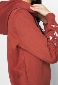 Röd hoodie i strukturerad tyg, med en dragsko på huvan och tryckta detaljer på ärmarna. Profilvy från sidan med händer som justerar huven.