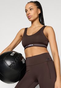 Sujetador deportivo marrón con tirantes anchos y el logo blanco de Adidas. La modelo sostiene una pelota medicinal negra. Mallas ajustadas marrones con una marca sutil de Adidas.