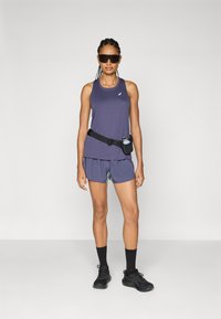 Camiseta de entrenamiento atlética morada y pantalones cortos con un diseño en capas, cinturón de correr negro con una botella de agua y gafas de sol oscuras. Calcetines y zapatos negros.