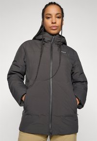 Vrouw met gevlochten haar die een zwarte gewatteerde hooded jacket draagt, met haar handen in haar zakken, tegen een effen lichte achtergrond.