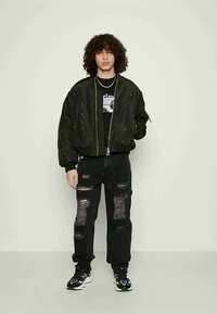 Μαύρο bomber jacket με ribbed μανίκια, φορεμένο πάνω από μαύρο graphic t-shirt. Συνδυασμένο με φθαρμένο μαύρο cargo παντελόνι και μαύρα sneakers.